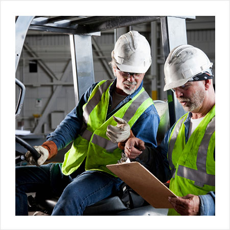 supervisor in safety vest with clip board reviewing information with a worker in a safety vest sitting on a forklift